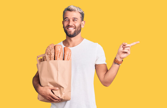 Young handsome blond man holding paper bag with bread smiling happy pointing with hand and finger to the side