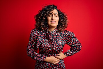 Young beautiful curly arab woman wearing casual floral dress standing over red background with hand on stomach because indigestion, painful illness feeling unwell. Ache concept.