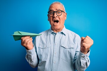 Middle age handsome hoary man holding paper airplane over isolated blue background screaming proud and celebrating victory and success very excited, cheering emotion