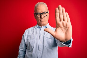 Naklejka premium Middle age handsome hoary man wearing casual striped shirt and glasses over red background doing stop sing with palm of the hand. Warning expression with negative and serious gesture on the face.