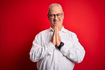 Middle age handsome hoary man wearing casual shirt and glasses over red background praying with hands together asking for forgiveness smiling confident.