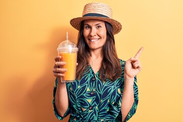 Young beautiful brunette woman wearing summer hat drinking glass of orange juice smiling happy pointing with hand and finger to the side