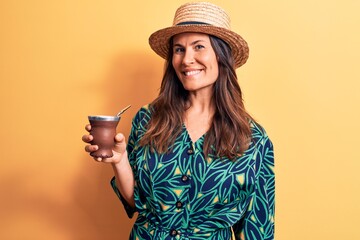 Young beautiful brunette woman wearing summer hat drinking cup of mate infusion beverage looking...