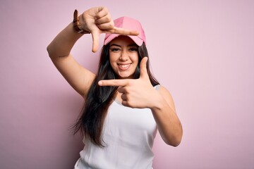 Obraz na płótnie Canvas Young brunette woman wearing casual sport cap over pink background smiling making frame with hands and fingers with happy face. Creativity and photography concept.