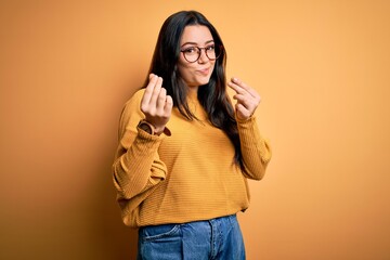 Young brunette woman wearing glasses and casual sweater over yellow isolated background doing money...