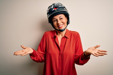 Middle age motorcyclist woman wearing motorcycle helmet over isolated white background clueless and confused expression with arms and hands raised. Doubt concept.