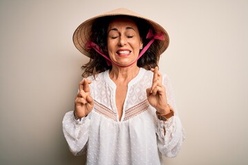 Middle age brunette woman wearing asian traditional conical hat over white background gesturing finger crossed smiling with hope and eyes closed. Luck and superstitious concept.