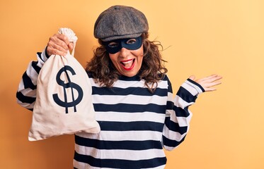 Middle age brunette burglar woman wearing mask and cap holding bag with dollar symbol celebrating...