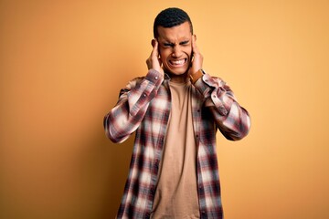 Young handsome african american man wearing casual shirt standing over yellow background covering ears with fingers with annoyed expression for the noise of loud music. Deaf concept.