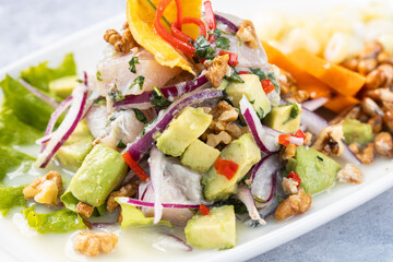 Close up shot of a white fish ceviche accompanied with avocado served on a white plate, isolated against a textured rustic gray wooden backdrop.