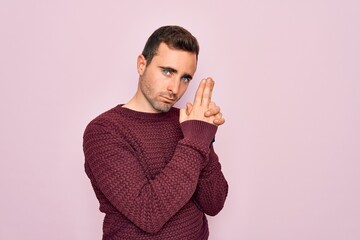 Young handsome man with blue eyes wearing casual sweater standing over pink background Holding symbolic gun with hand gesture, playing killing shooting weapons, angry face