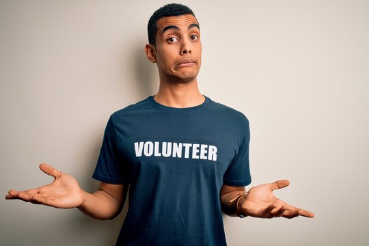 Young handsome african american man volunteering wearing t-shirt with volunteer message clueless and confused expression with arms and hands raised. Doubt concept.