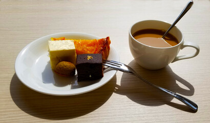 Close up image of small pieces of a peach pie, brownie, cookie, and a cheesecake on a small porcelain plate. A shiny fork is next to plate and all are served with a cup of milk coffee in a mug.
