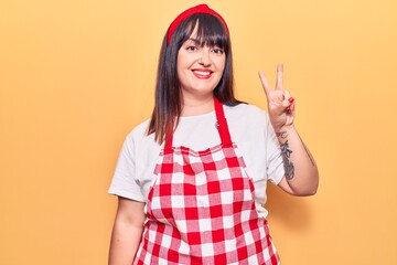 Young plus size woman wearing apron smiling with happy face winking at the camera doing victory sign. number two.