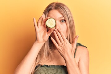 Young beautiful blonde woman holding slicie of cucumber covering mouth with hand, shocked and...
