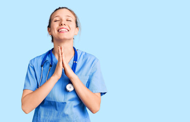 Young beautiful blonde woman wearing doctor uniform and stethoscope begging and praying with hands...