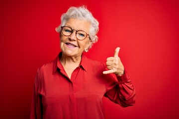 Senior beautiful grey-haired woman wearing casual shirt and glasses over red background smiling doing phone gesture with hand and fingers like talking on the telephone. Communicating concepts.