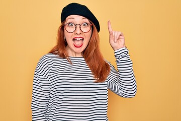 Beautiful redhead woman wearing striped t-shirt and french beret over yellow background pointing finger up with successful idea. Exited and happy. Number one.