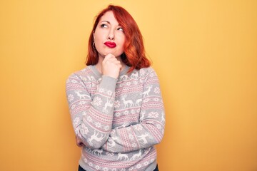 Beautiful redhead woman wearing christmas sweater with reindeer over yellow background thinking concentrated about doubt with finger on chin and looking up wondering