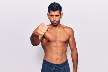 Young latin man standing shirtless angry and mad raising fist frustrated and furious while shouting with anger. rage and aggressive concept.