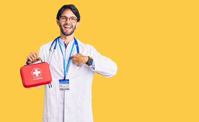 Handsome hispanic man wearing doctor coat holding first aid kit pointing finger to one self smiling...