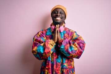 Young handsome african american man wearing colorful coat and cap over pink background praying with hands together asking for forgiveness smiling confident.
