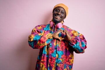 Young handsome african american man wearing colorful coat and cap over pink background smiling in love doing heart symbol shape with hands. Romantic concept.