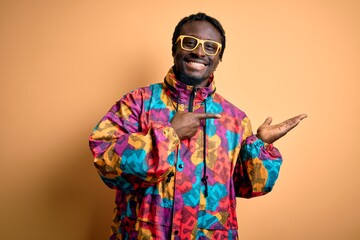 Handsome african american man wearing colorful coat and glasses over yellow background amazed and smiling to the camera while presenting with hand and pointing with finger.
