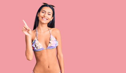 Young beautiful latin girl wearing bikini and sunglasses showing and pointing up with finger number one while smiling confident and happy.