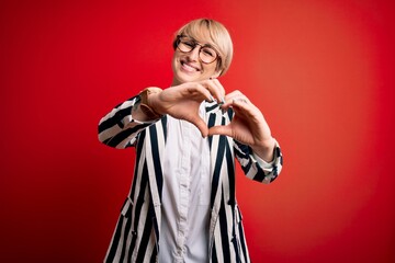 Blonde business woman with short hair wearing glasses and striped jacket over red background smiling in love doing heart symbol shape with hands. Romantic concept.