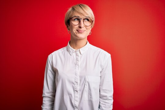 Young blonde business woman with short hair wearing glasses over red background smiling looking to the side and staring away thinking.
