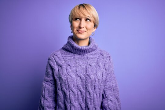 Young blonde woman with short hair wearing winter turtleneck sweater over purple background smiling looking to the side and staring away thinking.