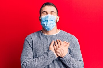 Young handsome man wearing medical mask smiling with hands on chest, eyes closed with grateful gesture on face. health concept.