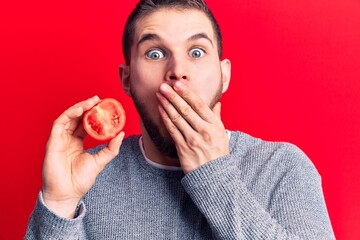 Young handsome man holding slice of tomato covering mouth with hand, shocked and afraid for mistake. surprised expression