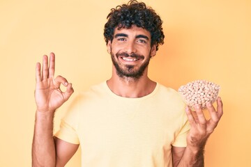 Handsome young man with curly hair and bear holding chickpeas bowl doing ok sign with fingers, smiling friendly gesturing excellent symbol