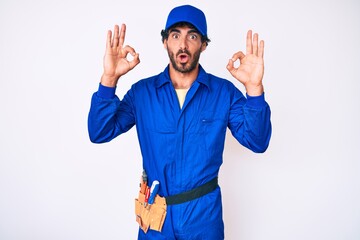 Handsome young man with curly hair and bear weaing handyman uniform looking surprised and shocked doing ok approval symbol with fingers. crazy expression