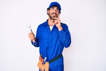 Handsome young man with curly hair and bear wearing builder jumpsuit uniform and holding wrench serious face thinking about question with hand on chin, thoughtful about confusing idea