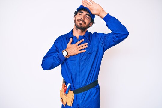 Handsome young man with curly hair and bear weaing handyman uniform touching forehead for illness and fever, flu and cold, virus sick