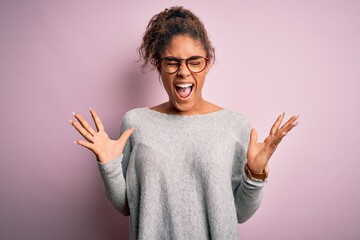 Young beautiful african american girl wearing sweater and glasses over pink background crazy and mad shouting and yelling with aggressive expression and arms raised. Frustration concept.