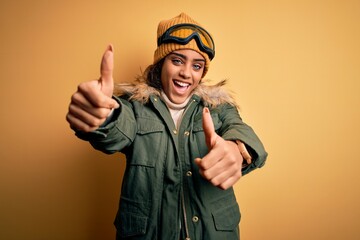 Young african american afro skier girl wearing snow sportswear and ski goggles approving doing positive gesture with hand, thumbs up smiling and happy for success. Winner gesture.