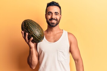 Young hispanic man holding melon looking positive and happy standing and smiling with a confident smile showing teeth
