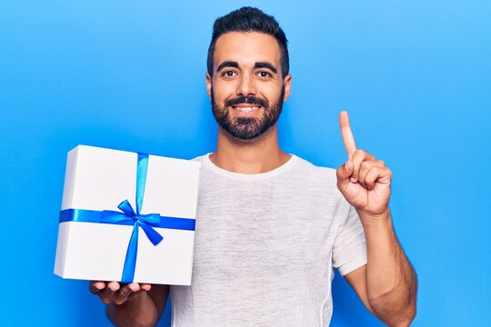 Young hispanic man holding gift smiling with an idea or question pointing finger with happy face, number one