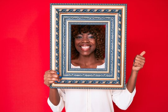 Young african american woman holding empty frame pointing thumb up to the side smiling happy with open mouth