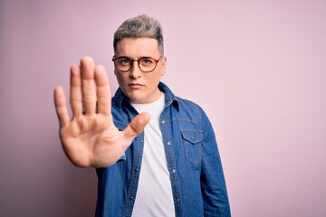 Young handsome modern man wearing glasses and denim jacket over pink isolated background doing stop sing with palm of the hand. Warning expression with negative and serious gesture on the face.