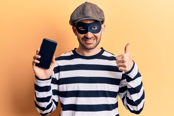 Young handsome bald man wearing burglar mask holding smartphone smiling happy and positive, thumb up doing excellent and approval sign