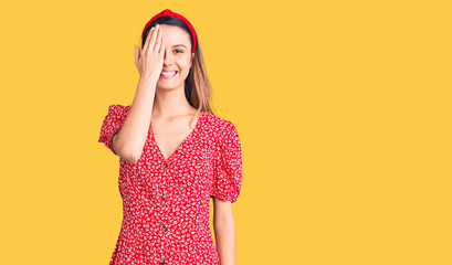 Young beautiful girl wearing dress and diadem covering one eye with hand, confident smile on face and surprise emotion.