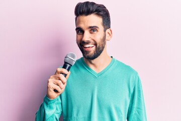 Young handsome man with beard singing song using microphone looking positive and happy standing and smiling with a confident smile showing teeth