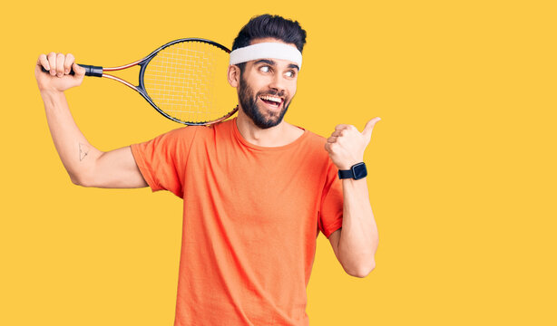 Young handsome man with beard playing tennis holding racket pointing thumb up to the side smiling happy with open mouth