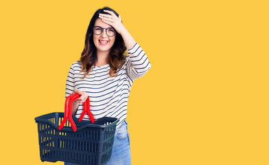Beautiful young brunette woman holding supermarket shopping basket stressed and frustrated with hand on head, surprised and angry face