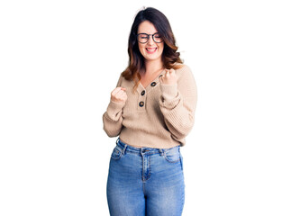 Beautiful young brunette woman wearing casual clothes and glasses celebrating surprised and amazed for success with arms raised and eyes closed. winner concept.